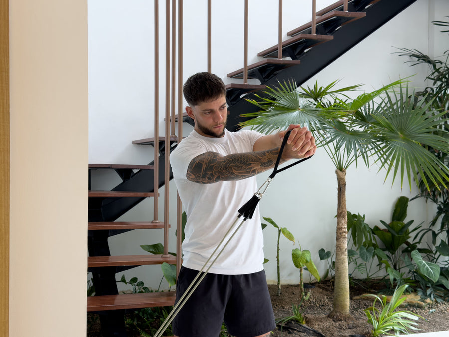 AEVO Fit resistance band woodchop exercise — man performing a low-to-high diagonal pull with both hands clasped, band anchored at floor level, staircase and tropical plants in background