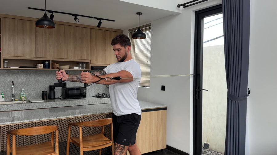 AEVO Fit resistance band chest fly — man performing a mid-height cable fly variation with arms extended forward and hands clasped, band anchored at shoulder height to the right, kitchen setting