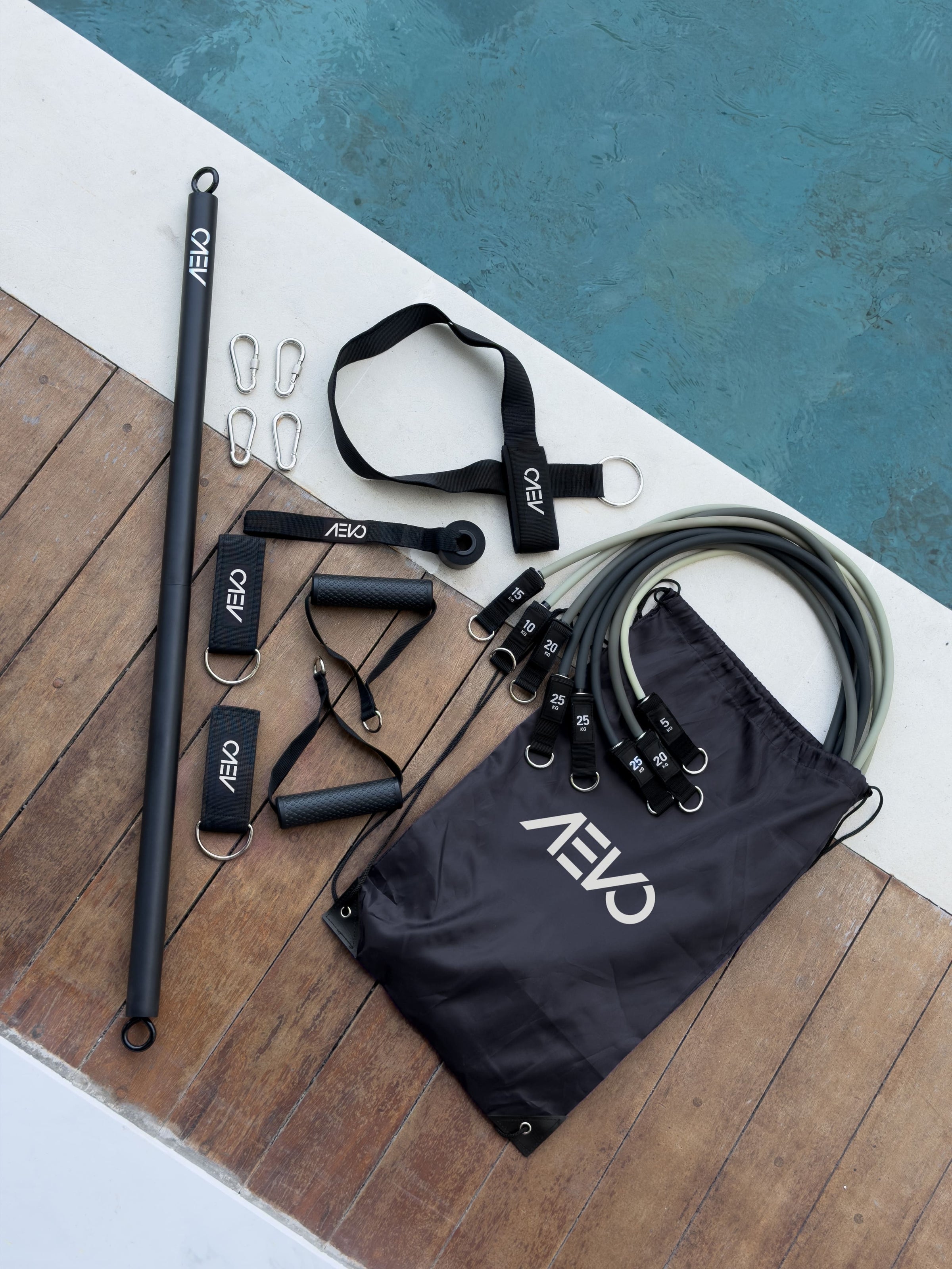 The AEVO Strength System laid out on decking beside a pool, showing the bar, resistance bands, handles and travel bag